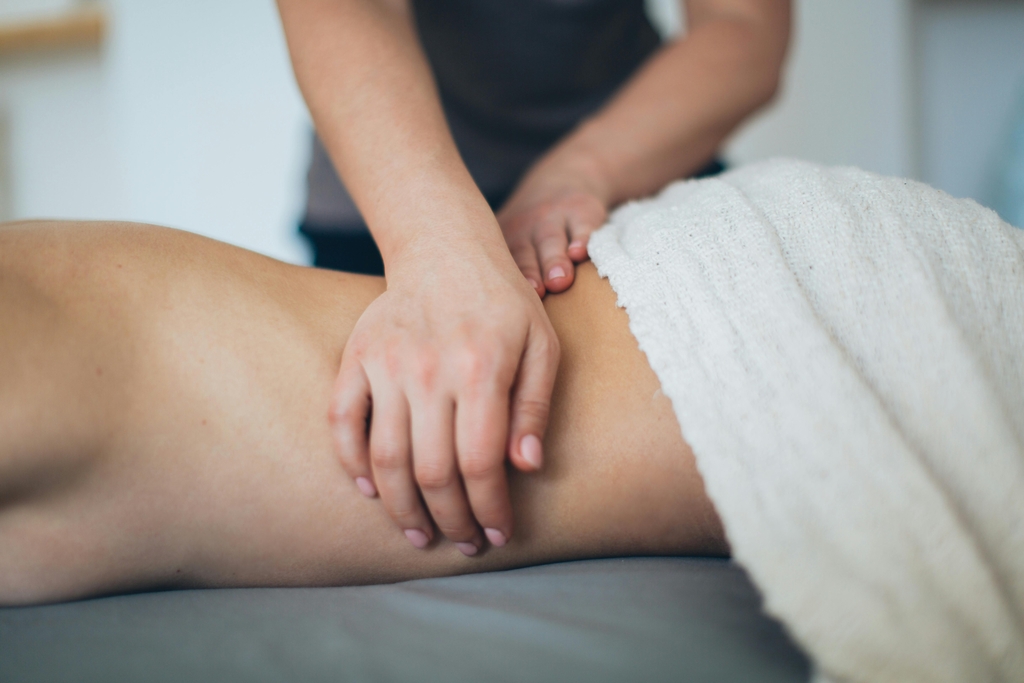 Massage therapist works on a client's lower back as the client lies on a massage table, while their lower body is covered with a white cloth. 