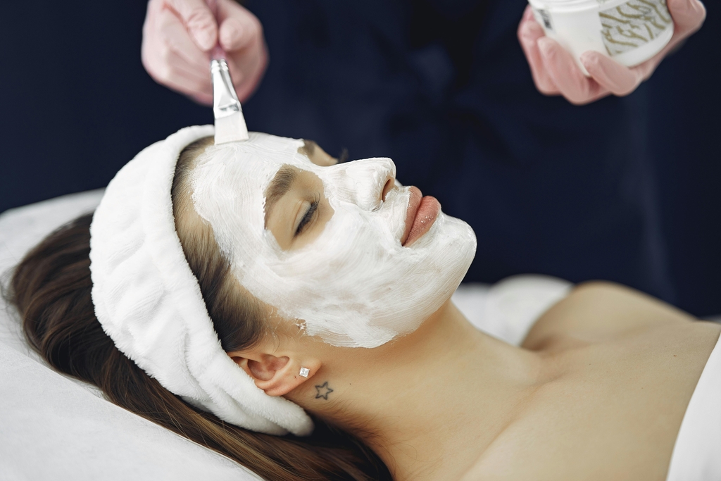 Woman with a white headband relaxes on a spa bed while an esthetician applies a white face mask with a soft brush.