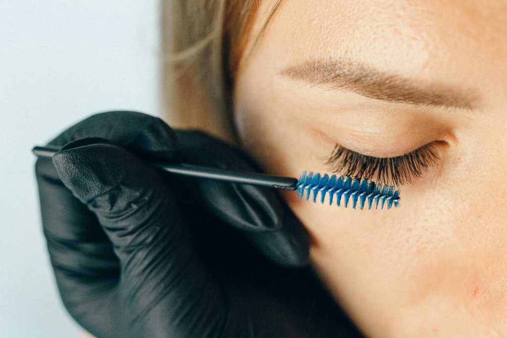 Esthetician wearing black medical gloves uses a blue eyelash brush on a client's eyelashes. 